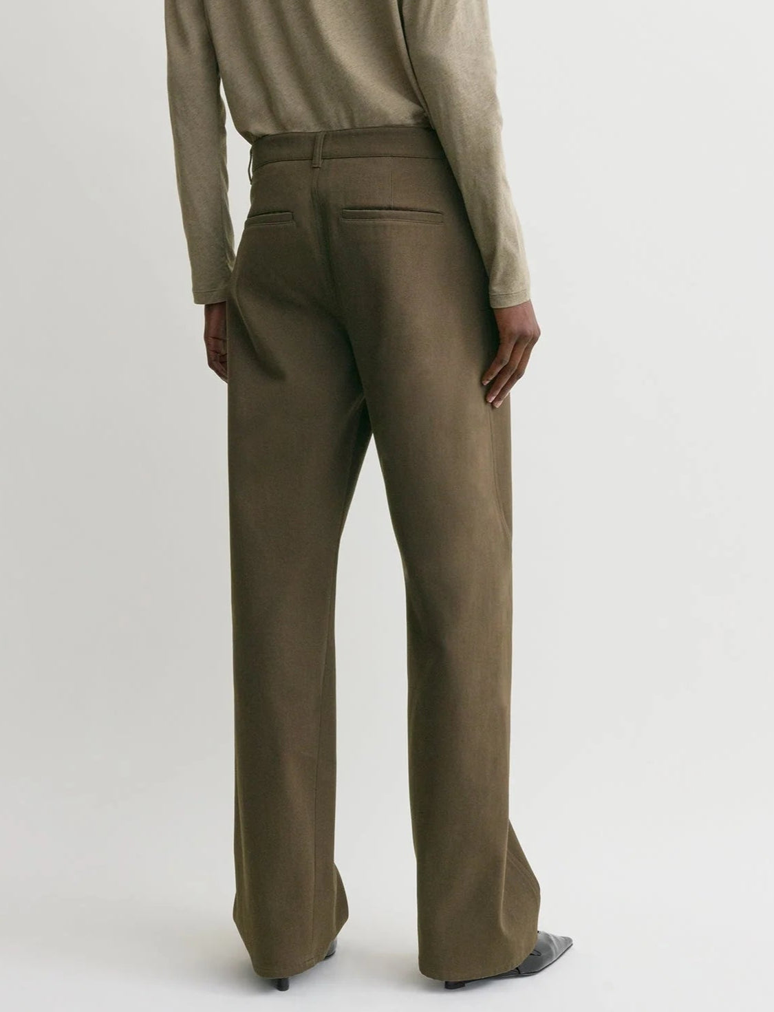 Model wears Toteme's Twisted Tailored Trousers in olive-green cotton, featuring a straight-leg cut and styled with a long-sleeve top, photographed from the back against a plain light background.