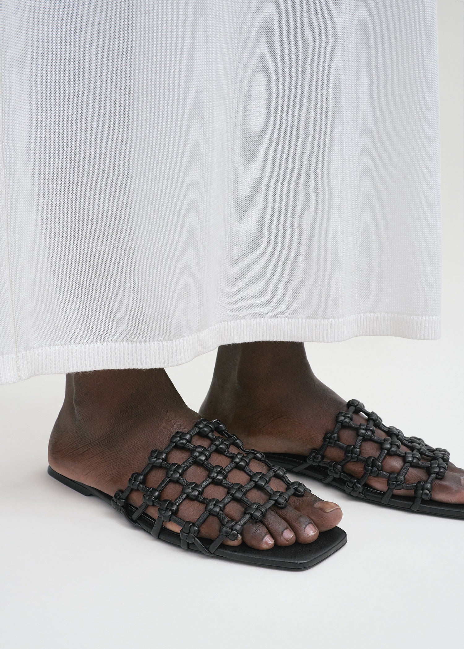 A person stands on a light surface wearing a long white garment and Toteme Braided Leather Bandeau Slides.