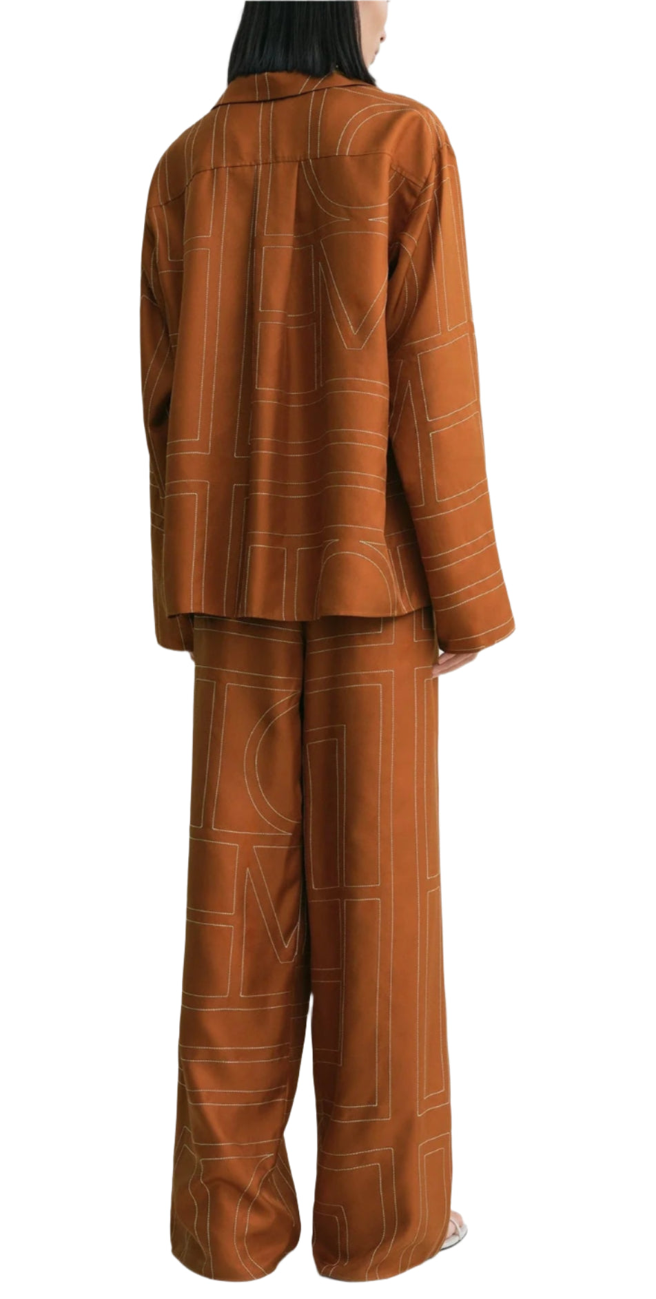 A person shown from the back wears Toteme's Monogram Silk PJ Top, a loose-fitting, rust-colored top with a geometric line pattern and silk twill texture.