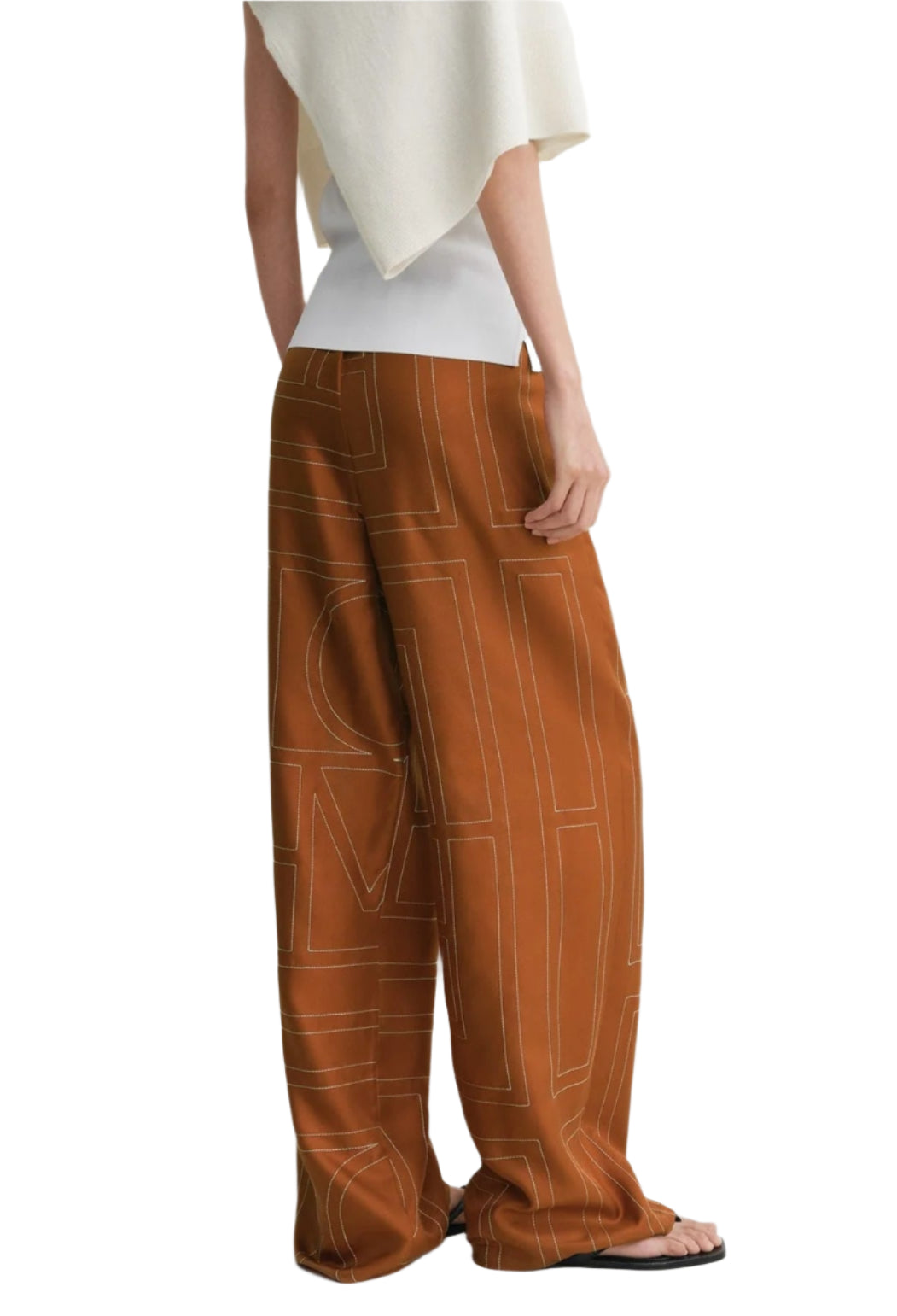 A person wearing Toteme Monogram Silk PJ Bottoms, a white top, and black sandals, photographed from the side.