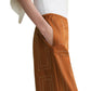 A person wears a white top with wide sleeves and rust-colored Toteme Monogram Silk PJ Bottoms featuring geometric stitching, standing with one hand in their pocket.