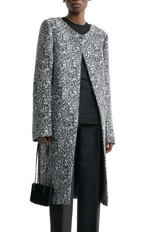 A person wears the Toteme Collarless Floral Coat in black and white over a black top and pants, accessorized with a small black handbag—a chic outfit for any occasion.