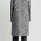 A person stands with their back to the camera in a Toteme Collarless Floral Coat—black-and-white jacquard over black pants and shoes—creating an effortlessly chic occasion look against a plain background.