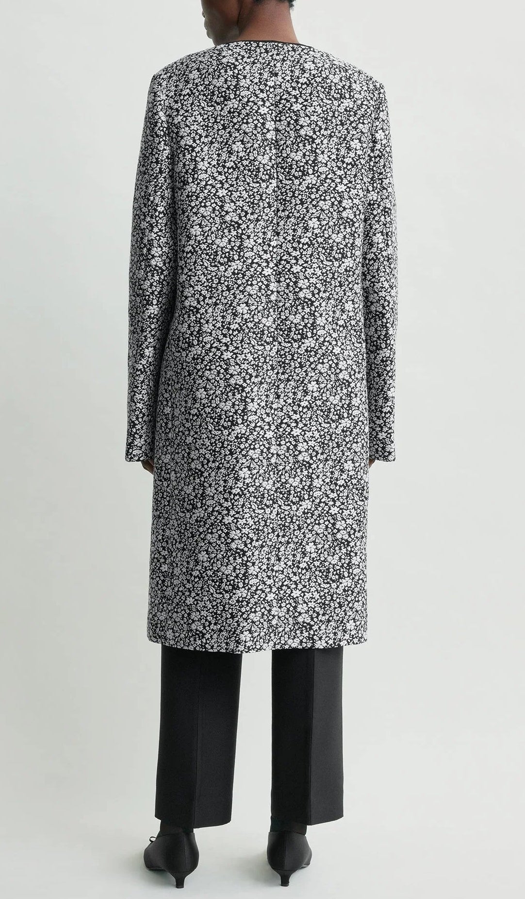 A person stands with their back to the camera in a Toteme Collarless Floral Coat—black-and-white jacquard over black pants and shoes—creating an effortlessly chic occasion look against a plain background.