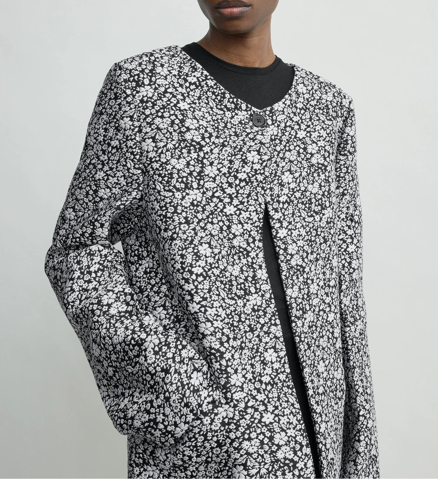 Model wears Toteme’s Collarless Floral Coat—black and white jacquard with round neckline, large button, and pockets—against a plain light background. Ideal for an elevated occasion look.