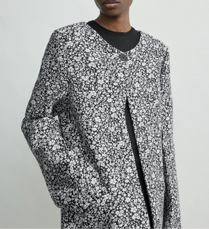 Model wears Toteme’s Collarless Floral Coat—black and white jacquard with round neckline, large button, and pockets—against a plain light background. Ideal for an elevated occasion look.