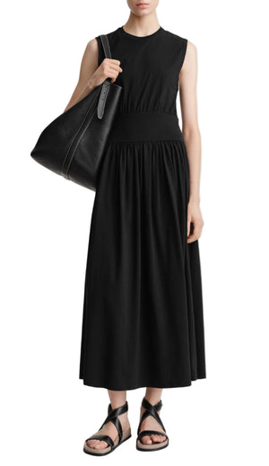 A woman wears Toteme’s Sleeveless Cotton Tee Dress in black, styled with black strappy sandals and a large black tote bag over her shoulder.