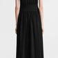 A person stands facing away, wearing Toteme's Sleeveless Cotton Tee Dress in black organic-cotton jersey with a wide waistband, paired with black sandals.