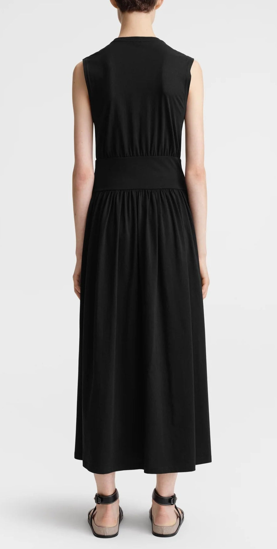 A person stands facing away, wearing Toteme's Sleeveless Cotton Tee Dress in black organic-cotton jersey with a wide waistband, paired with black sandals.