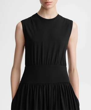 A person wearing the Toteme Sleeveless Cotton Tee Dress, a black organic-cotton jersey dress with a fitted waistband and gathered skirt, is photographed from the shoulders to just above the knees against a plain background.
