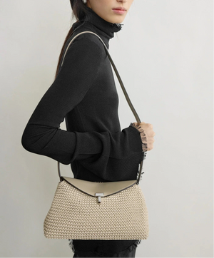 Someone wearing a black long-sleeve top holds the Toteme T-Lock Woven Leather Clutch, a beige bag with monochrome braiding and a flap with clasp closure, against a plain light background.