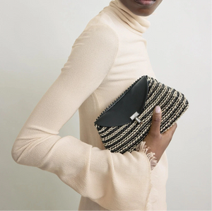 A person in a long-sleeved, textured beige dress holds Toteme's Mini T-Lock Woven Clutch against a plain light background.