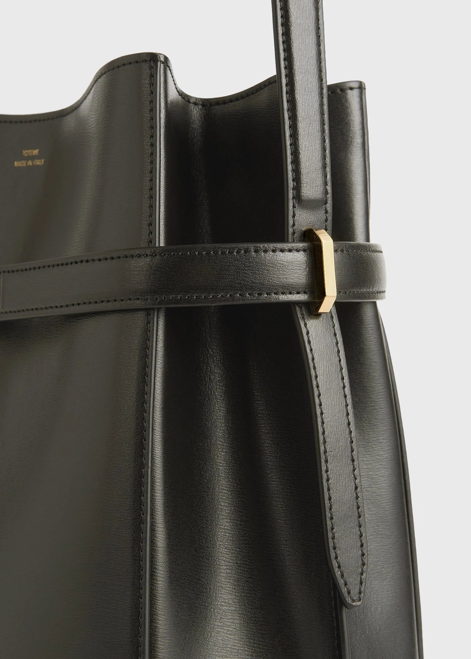 Close-up of the Toteme Belted Bucket Bag in black Palmellato leather, featuring a gold rectangular buckle and stitched strap detailing.