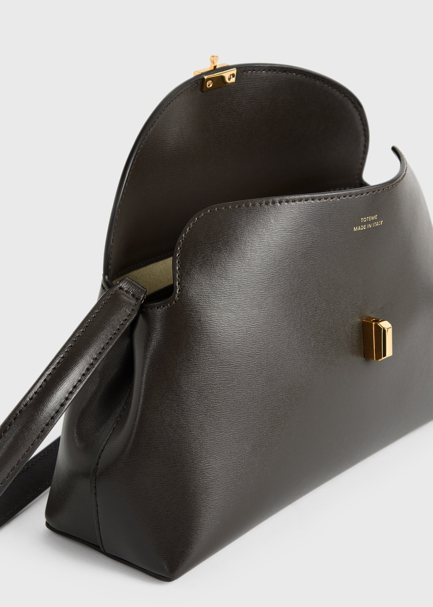 The Toteme T-Lock Clutch is a black Palmellato leather crossbody bag featuring a gold T-Lock clasp, shown partially open to reveal its interior.