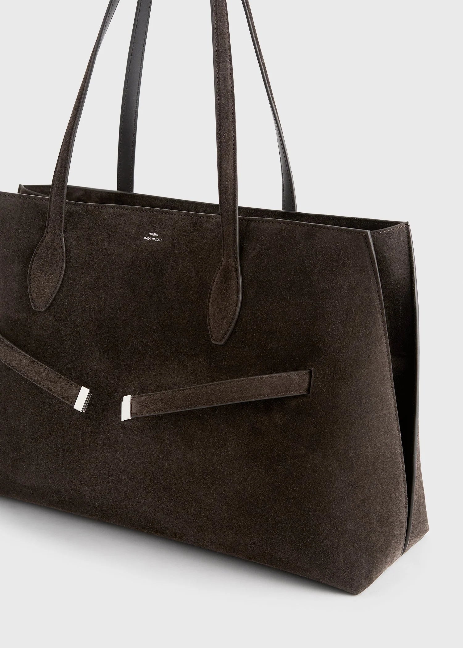 The Suede Lounge Tote by Toteme is a large, dark brown Italian suede bag with two top handles and a minimalist design, featuring a subtle metallic accent—ideal for everyday use.