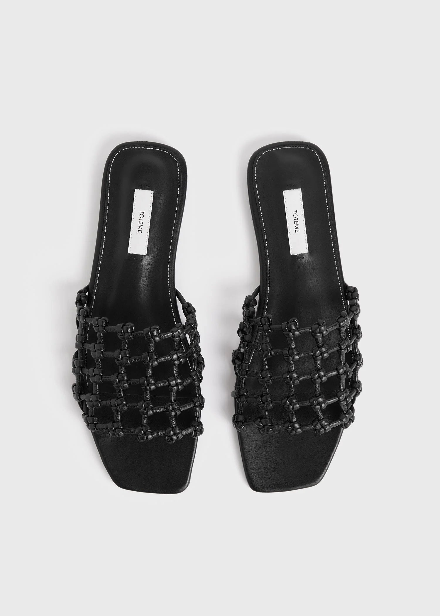 A pair of Toteme Braided Leather Bandeau Slides in black with flat soles, open toes, braided knotted leather straps, and square toes, shown on a white background.