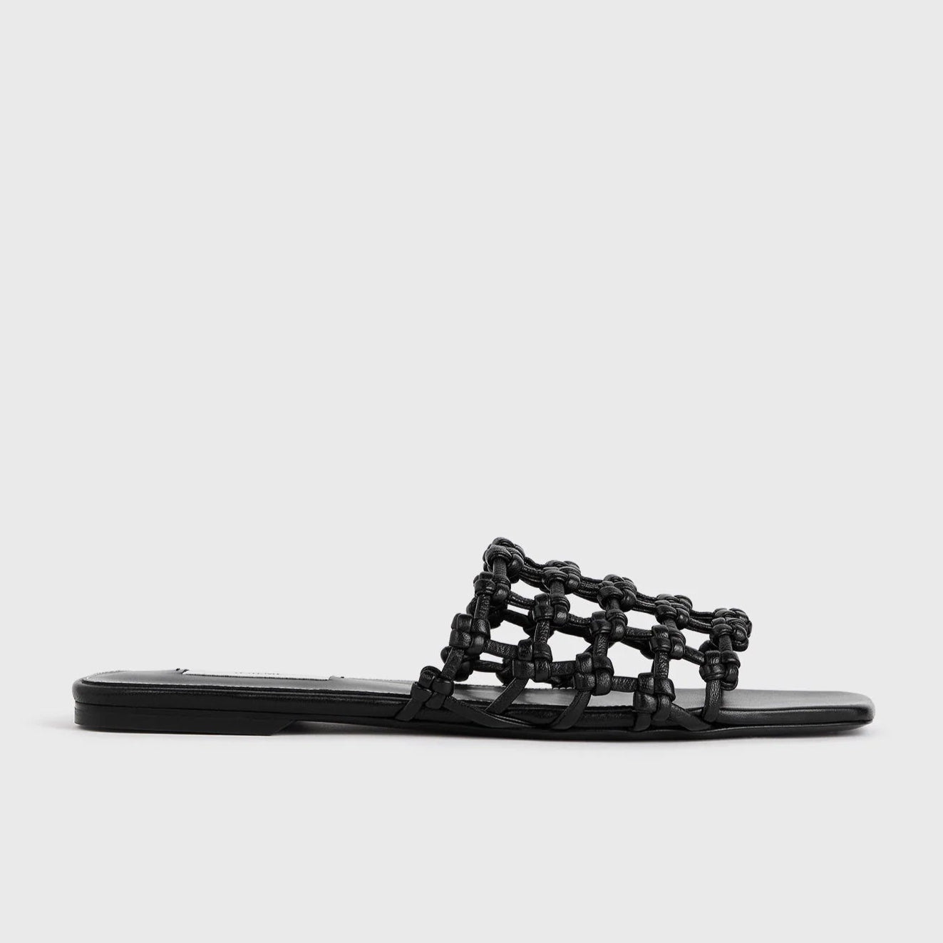 The Braided Leather Bandeau Slides by Toteme feature a black, flat design with an open, braided leather upper and thin sole, shown on a light gray background—an elegant update to classic Toteme slides.