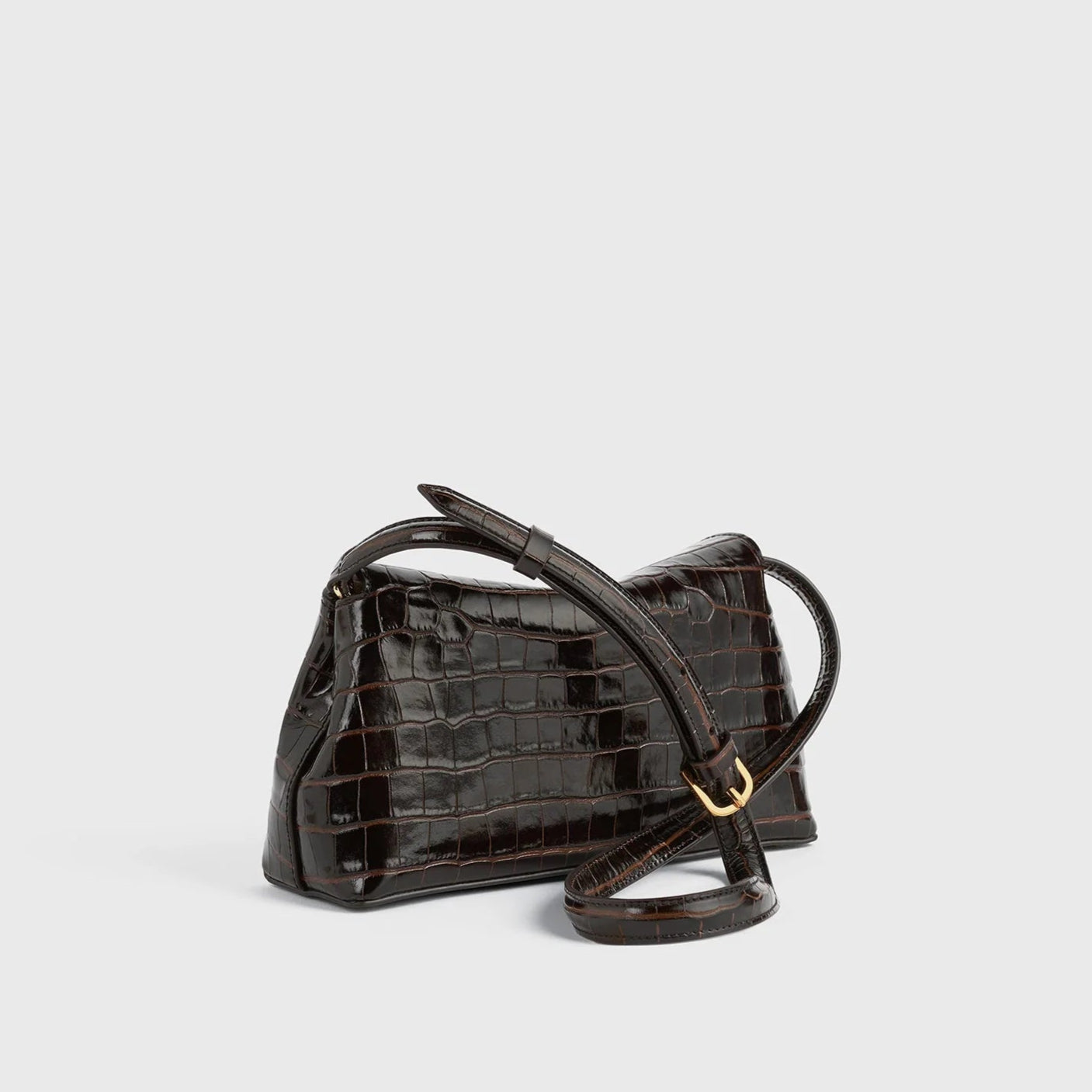 The Toteme T-Lock Embossed Croc Clutch is a small, structured bag in dark brown crocodile-effect leather with a long adjustable strap and gold buckle, shown against a plain light background.
