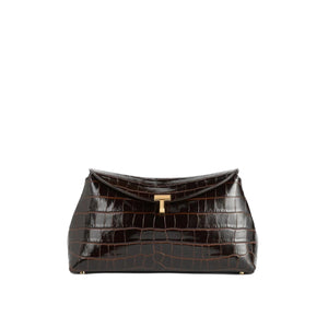 The Toteme T-Lock Embossed Croc Clutch in dark brown features crocodile-effect leather and a gold-tone T-Lock clasp, shown against a plain white background.