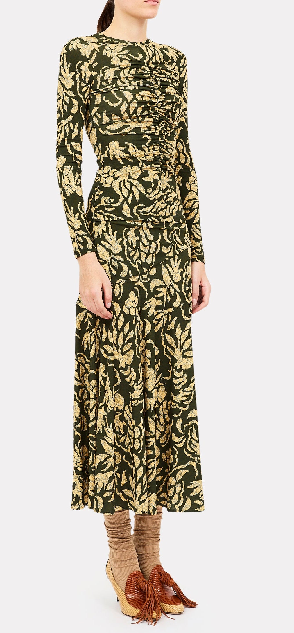 A person wearing a long-sleeve, ruched olive green and beige floral dress paired with the Ulla Johnson Fiona Jersey Skirt, tan socks, and beige shoes with large brown tassels stands against a plain background.
