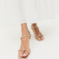 A woman wearing the Loeffler Randall Triana Mid Heel Sandal, featuring metallic strappy details and square toes, pairs it with a white flowy skirt while standing against a white background. The mid-heel sandal perfectly complements her elegant look.