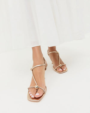 A woman wearing the Loeffler Randall Triana Mid Heel Sandal, featuring metallic strappy details and square toes, pairs it with a white flowy skirt while standing against a white background. The mid-heel sandal perfectly complements her elegant look.
