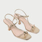 Introducing the Triana Mid Heel Sandal by Loeffler Randall—a pair of gold, open-toe strappy sandals with slim ankle straps, square toes, and a decorative metallic bead detail at the front. These chic mid-heel sandals effortlessly combine elegance and comfort.