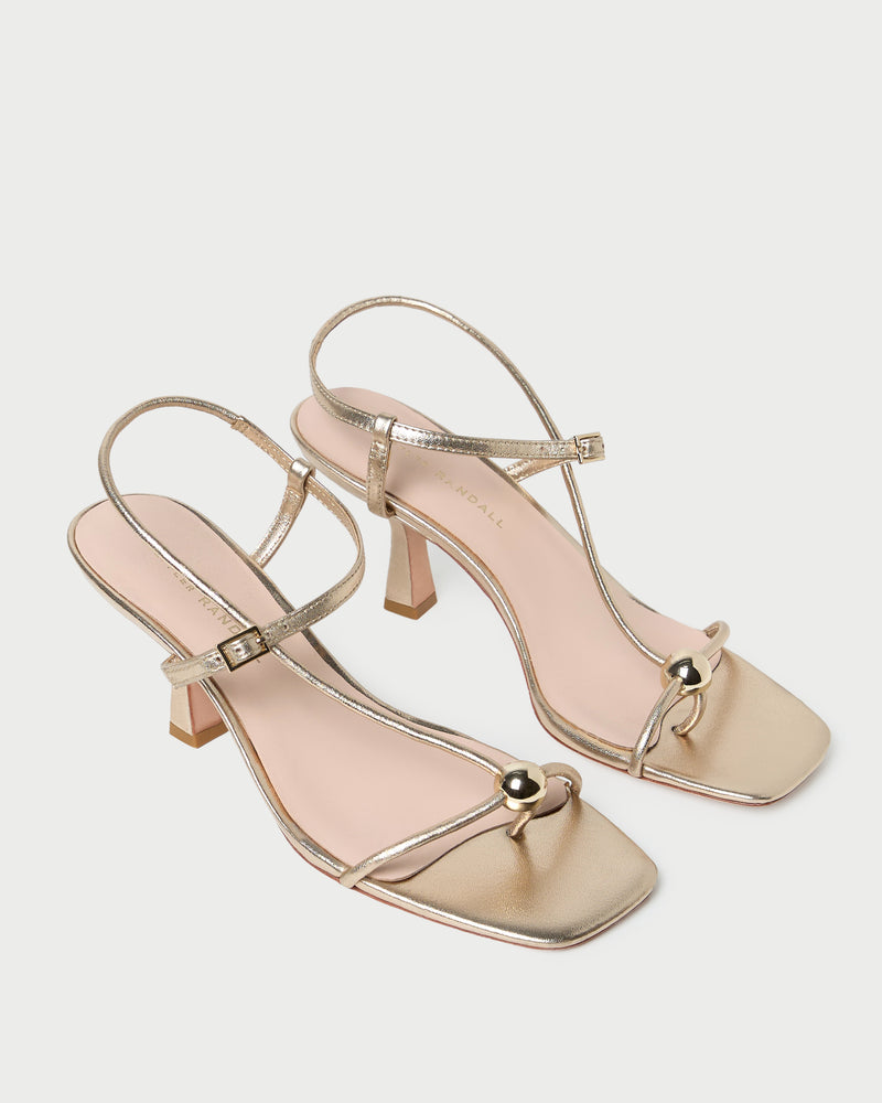 Introducing the Triana Mid Heel Sandal by Loeffler Randall—a pair of gold, open-toe strappy sandals with slim ankle straps, square toes, and a decorative metallic bead detail at the front. These chic mid-heel sandals effortlessly combine elegance and comfort.