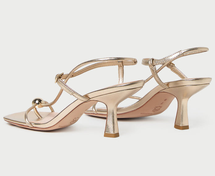 The Loeffler Randall Triana Mid Heel Sandal is featured here in a striking metallic gold finish, showcasing delicate straps, a square toe, and a chic mid-height heel against a plain light background. This elegant design brings a contemporary edge to the timeless strappy sandal silhouette.