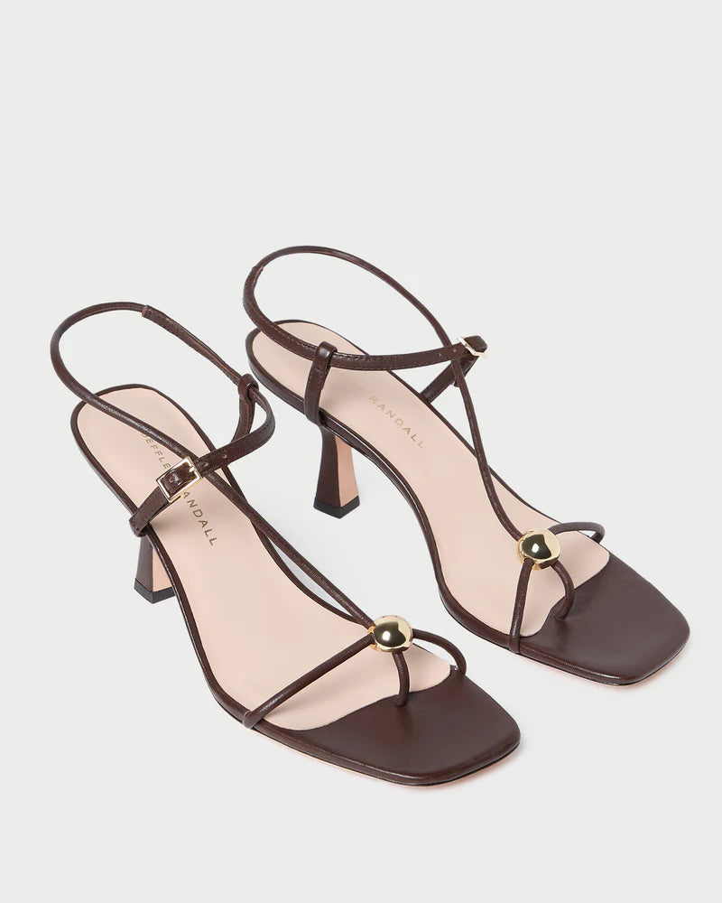 The Loeffler Randall Triana Mid Heel Sandal features brown nappa leather, strappy design, square toes, and gold round accents on the front straps, shown against a white background.