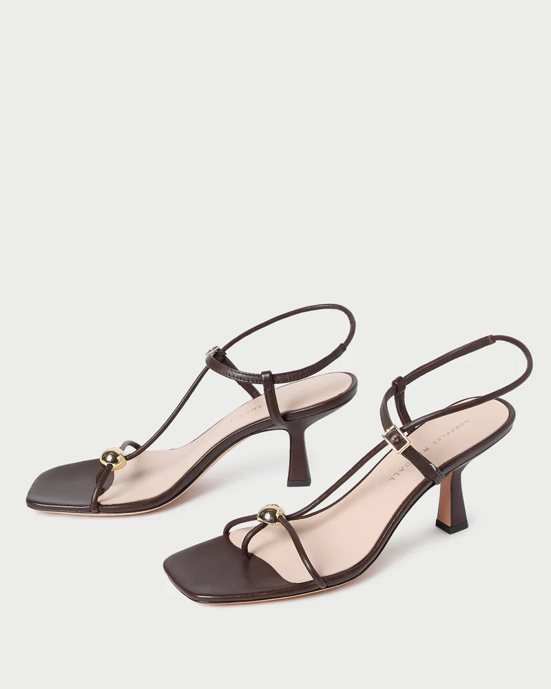 The Loeffler Randall Triana Mid Heel Sandal features brown nappa leather, square toes, thin straps with ankle buckles, and a gold ornament on each front strap, shown against a white background.