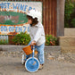 A child wearing a helmet stands next to the Banwood Kids’ Trike Vintage, a blue tricycle with a wicker basket, outdoors near a rustic sign and greenery.