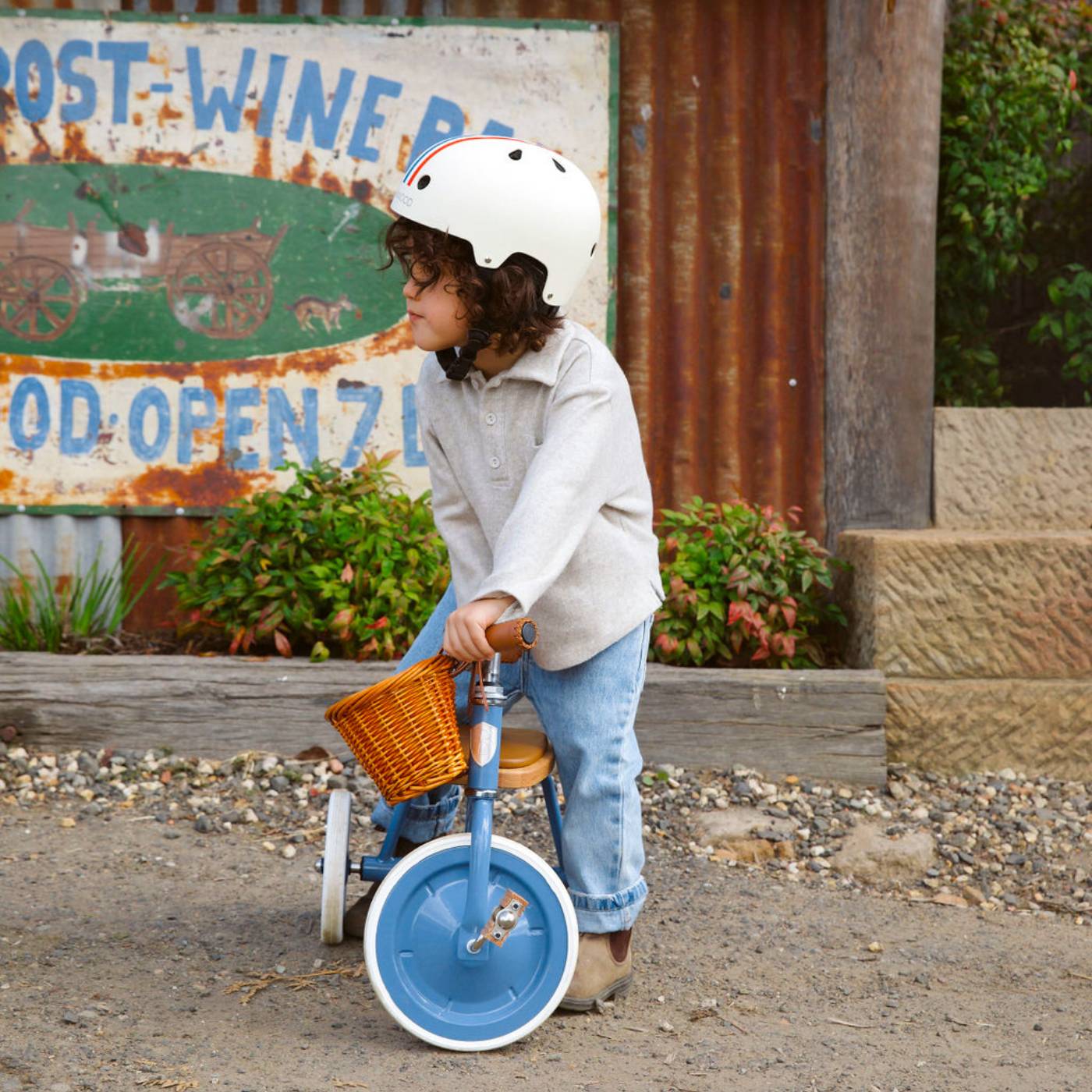 A child wearing a helmet stands next to the Banwood Kids’ Trike Vintage, a blue tricycle with a wicker basket, outdoors near a rustic sign and greenery.