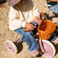 A child in a cream sweater and jeans sits on a Banwood Kids’ Trike Vintage, holding a toy camera, with a wicker basket attached to the handlebars, on a gravel surface.