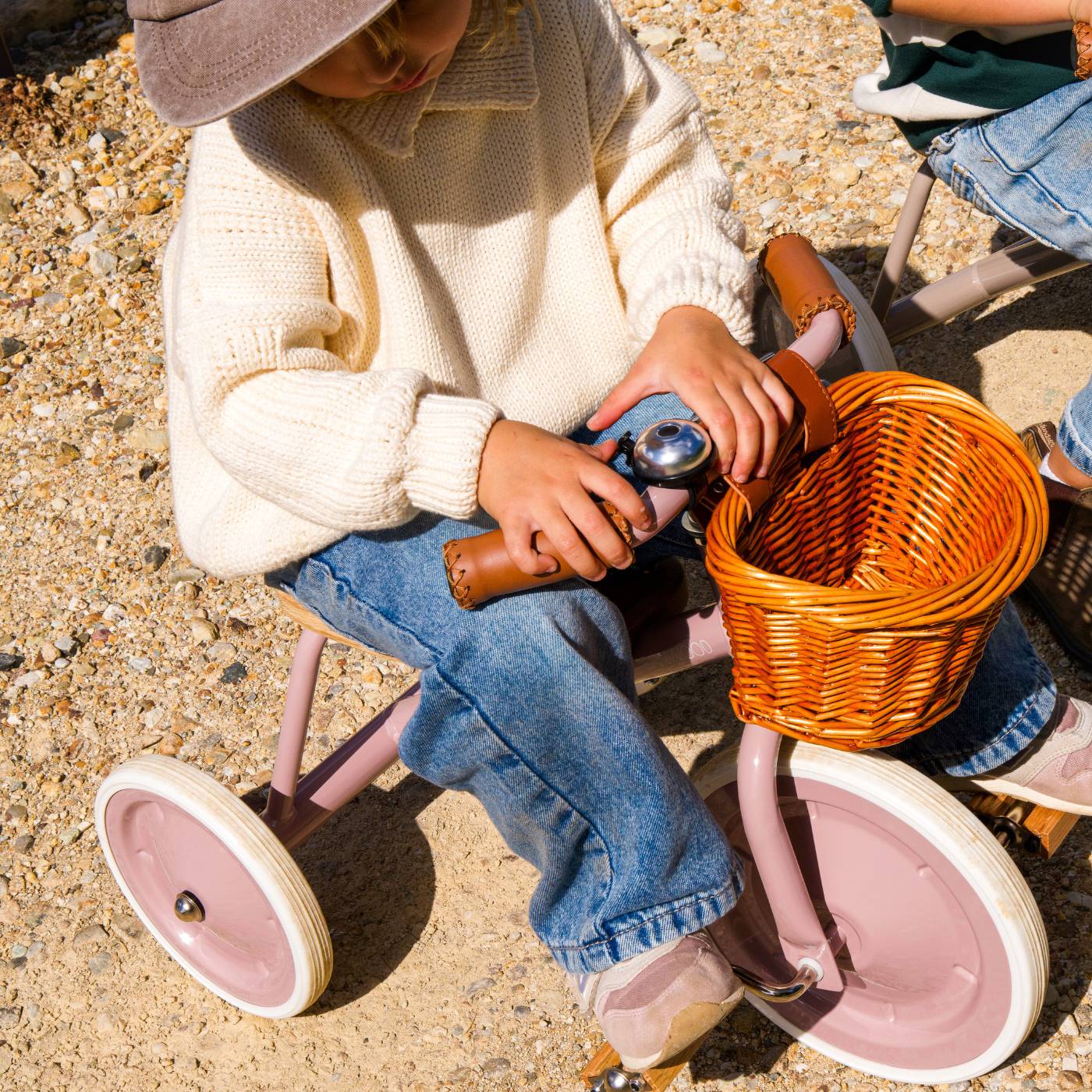 A child in a cream sweater and jeans sits on a Banwood Kids’ Trike Vintage, holding a toy camera, with a wicker basket attached to the handlebars, on a gravel surface.