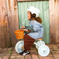 A young child wearing a helmet and denim jacket rides a Banwood Kids’ Trike with an orange basket, set in front of a rustic wooden wall.