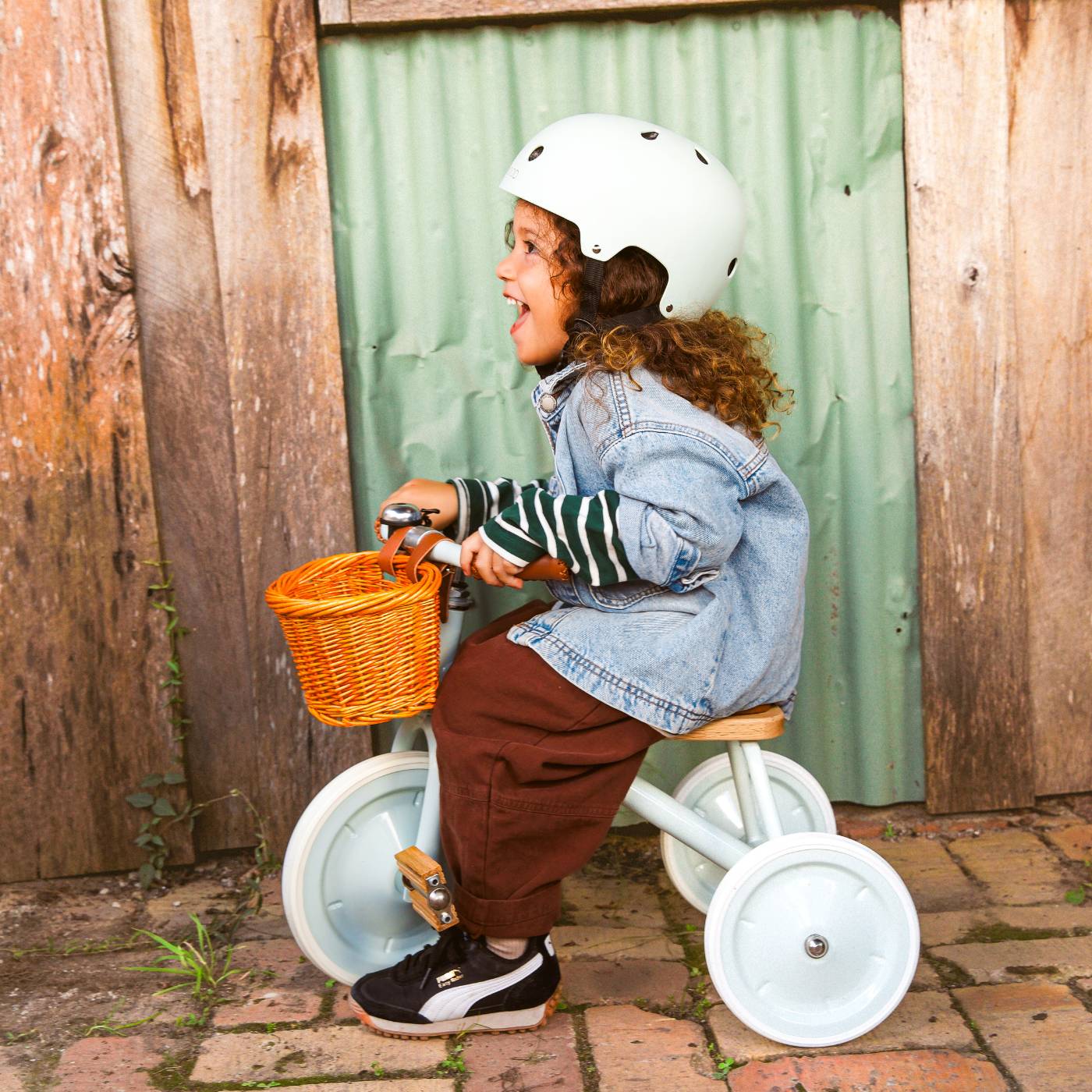 A young child wearing a helmet and denim jacket rides a Banwood Kids’ Trike with an orange basket, set in front of a rustic wooden wall.