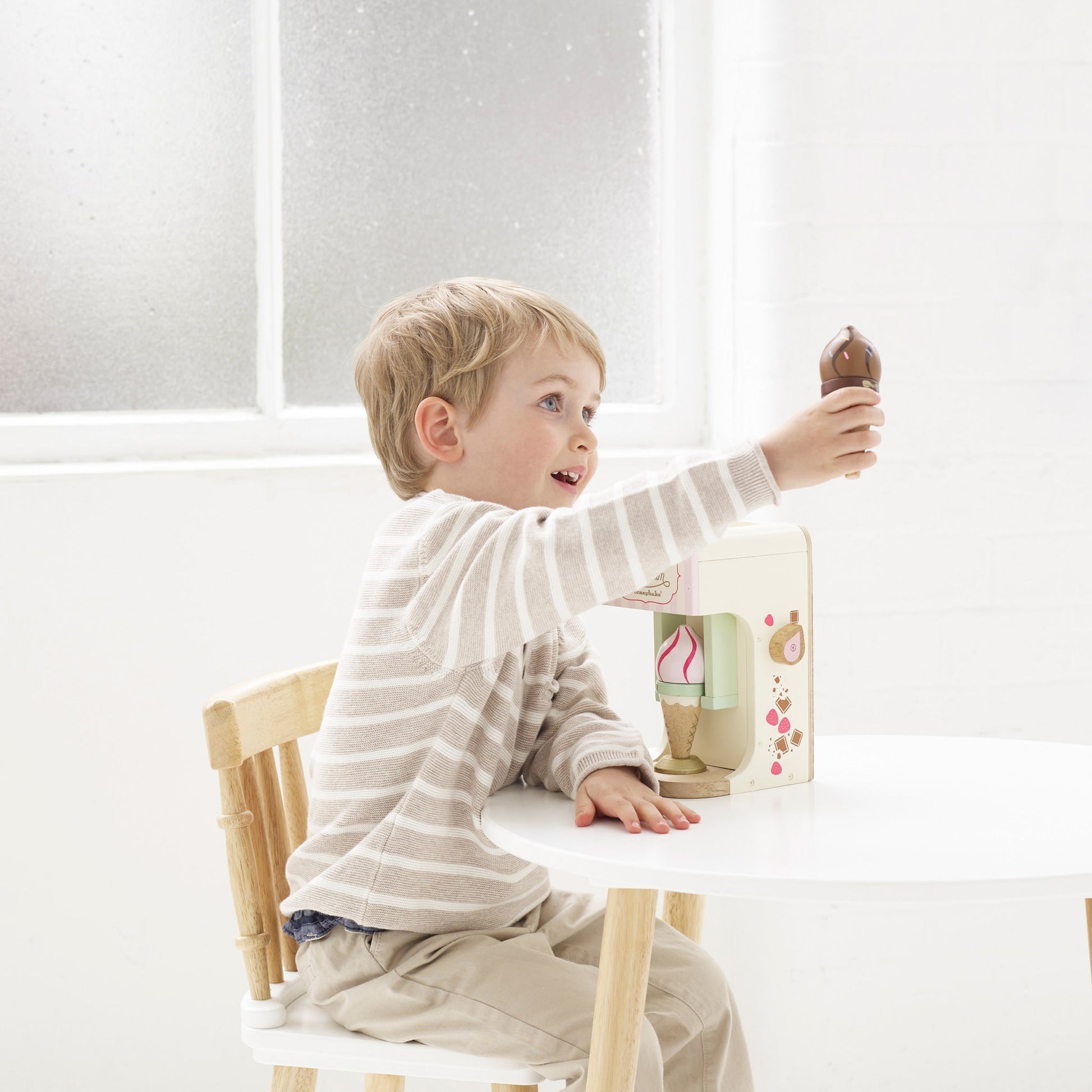 A young boy reaching for a Le Toy Van Ice Cream Machine at a wooden table with a decorated box.