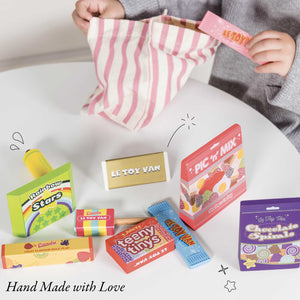 A child packs a striped cloth bag with assorted colorful toy candy boxes on a white table. The items, part of the Le Toy Van Sweet & Candy Set from sustainable rubberwood, include "LE TOY VAN," "PIC 'N' MIX," "Rainbow Stars," "teeny tins," and "Chocolate Sprinkles.