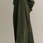 A person wearing TWP's Jenny's Coated Linen Gown, a green button-up dress with a point collar made from coated viscose linen, and black sandals stands against a plain background with hands behind back.
