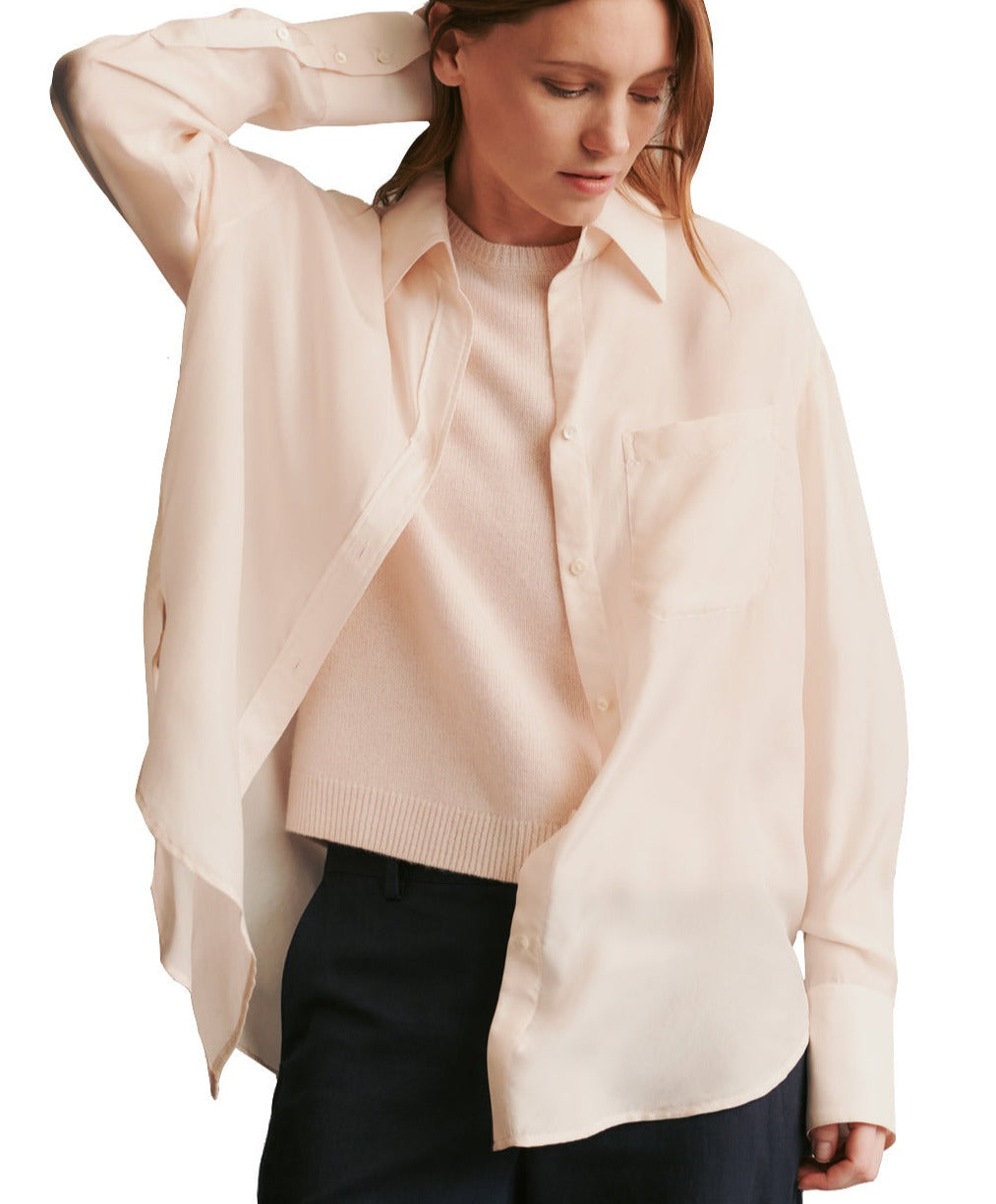 A person wearing an oversized, light pink TWP Big Joe Silk Top layered over a textured pink sweater, paired with dark pants, poses with one hand on their head.