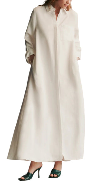 A person wears the TWP Jennys Coated Linen Gown, a long, oversized white dress with long sleeves and a collar, paired with dark green heeled sandals.