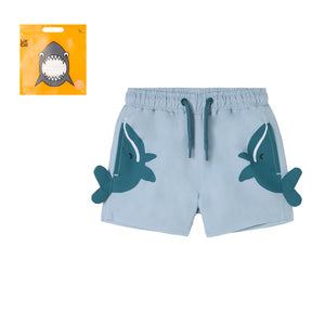 Stella McCartney Kids' Baby Boys' Whale Swim Shorts are light blue, quick-drying, with playful shark designs on the sides, and come beside bold orange packaging featuring a cartoon shark face.