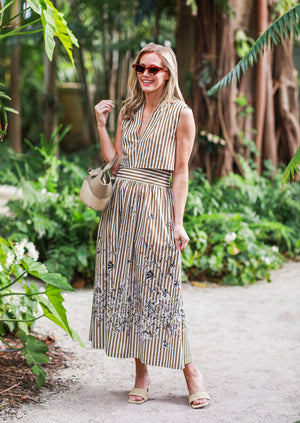 A woman stands on a garden path wearing the Lela Rose Collared V-Neck Dress, paired with sunglasses, sandals, and holding a woven handbag.