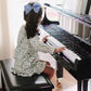 A young child wearing the Timo & Violet Girls’ Sunday Morning Dress with a blue bow in her hair sits on a bench, facing away, and plays a black piano in a sunlit room.