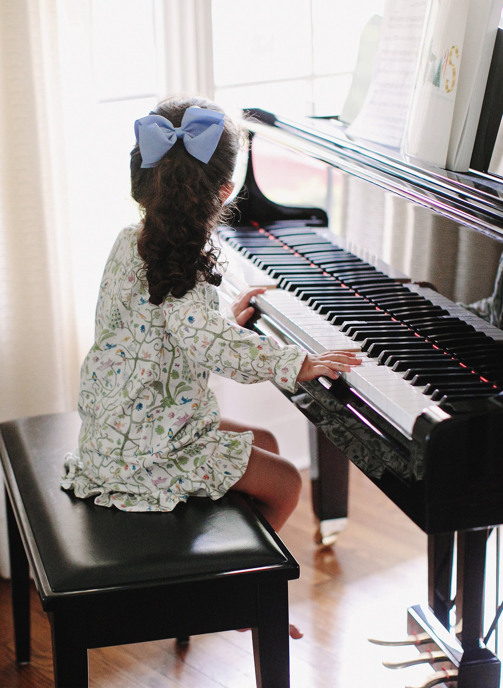 A young child wearing the Timo & Violet Girls’ Sunday Morning Dress with a blue bow in her hair sits on a bench, facing away, and plays a black piano in a sunlit room.