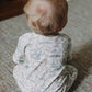 A young child with light hair sits on a textured rug, facing away from the camera and wearing the Timo & Violet Baby Walker Terry Onesie.