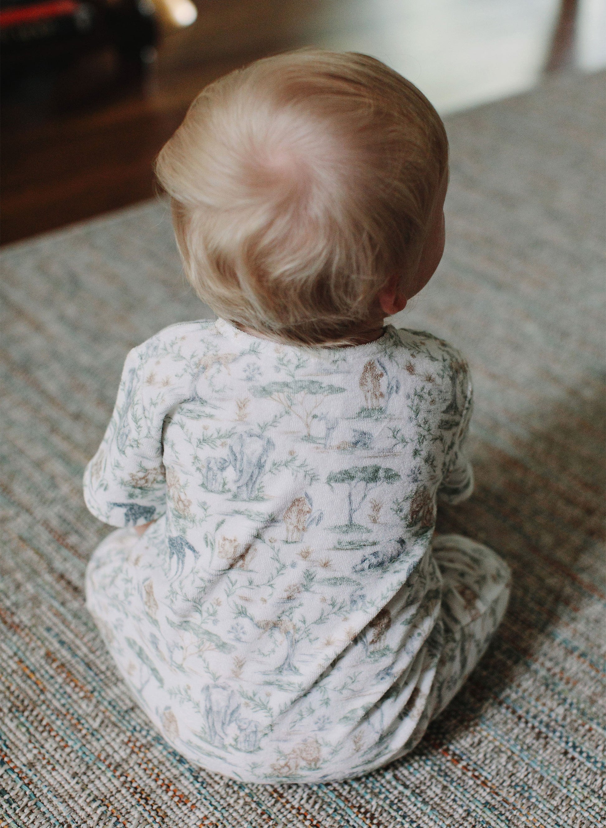 A young child with light hair sits on a textured rug, facing away from the camera and wearing the Timo & Violet Baby Walker Terry Onesie.
