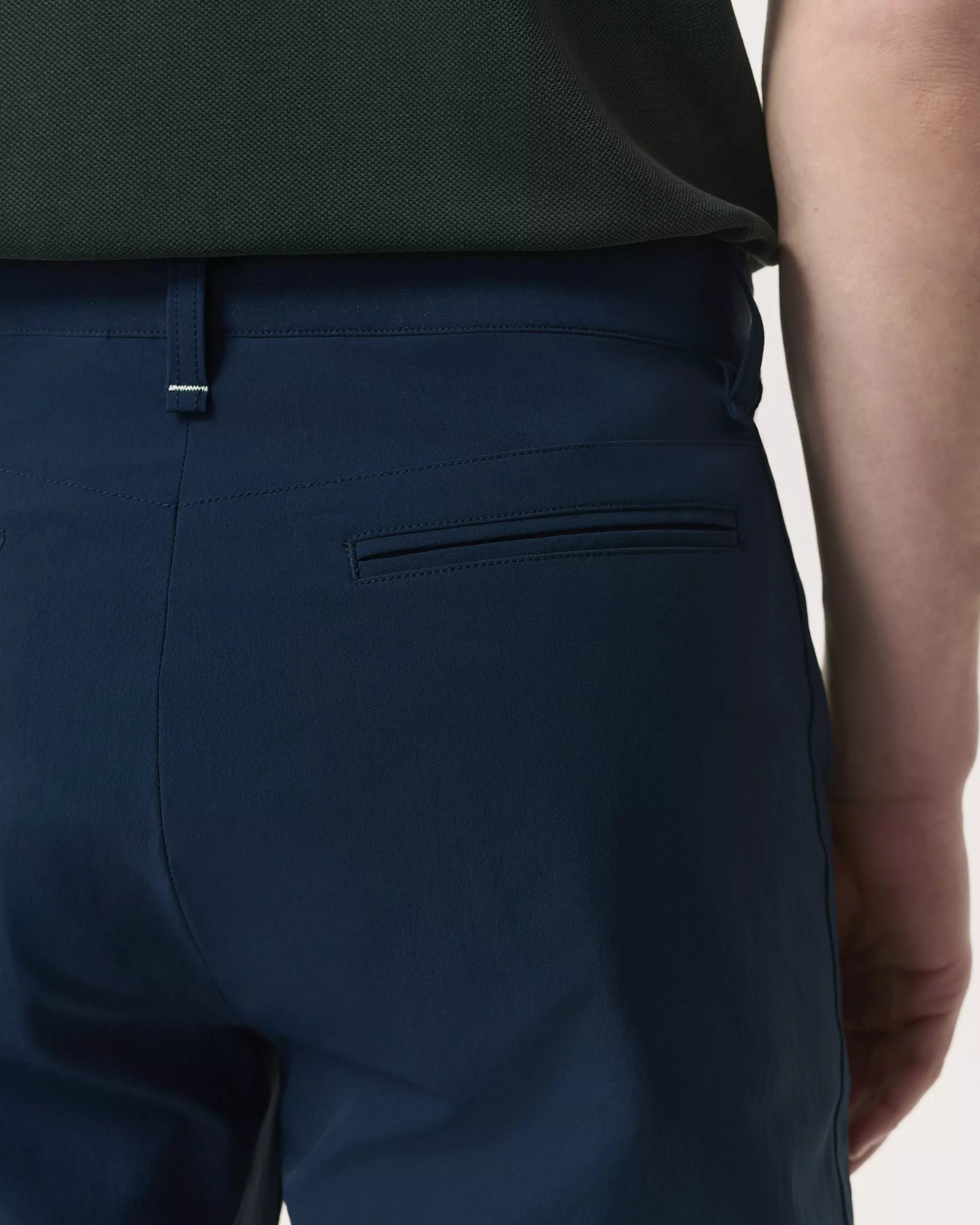 Close-up of a person wearing Rag & Bone's Tapered Nylon-Blend Trouser in dark blue, paired with a dark green shirt, showcasing the waistband, belt loop, and back pocket details on these streamlined pants.