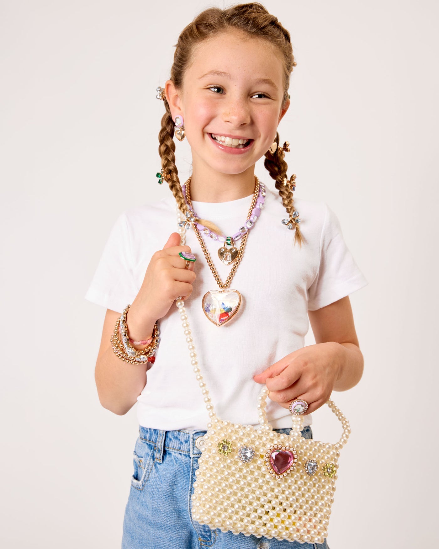 Smiling girl with braided hair holds the Super Smalls Pearl of the Party Handbag, decorated with gems—a playful dress-up bag that pairs perfectly with a white t-shirt, blue jeans, and colorful jewelry.